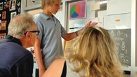 A young boy is standing in front of a board in a classroom, pointing to something and talking to two adults.