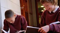 Two boys sitting in the hallway writing in their notebooks, talking, and smiling