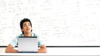A student works at a desk in front of a whiteboard covered with equations.
