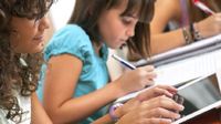 A girl writing in a binder and sitting next to her, a girl on a tablet