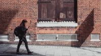 A young woman is walking on the sidewalk past a brick building with her backpack on, looking at her cellphone.