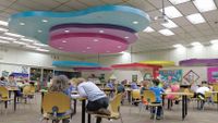 Young students are working at tables in a large multipurpose room with large colorful-shaped decorations hanging from the ceiling.