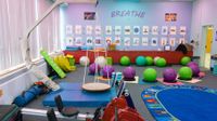 A colorful classroom with light purple walls, blue and purple cloths covering the ceiling lights, a swing set, bouncy balls to sit on, and rowing exercise equipment.