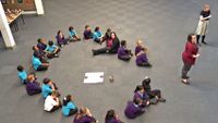 22 primary students and one teacher sitting in circle on the floor in assembly with two teachers standing on the side