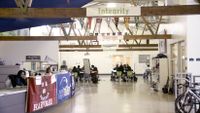 A multipurpose room filled with college banners, students studying at tables, cubbies against one wall, and an open office with two adults sitting behind a desk counter.