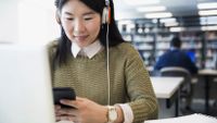 A female student listens to music and checks her phone while doing schoolwork.