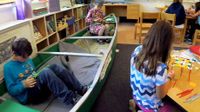 Kids working independently sitting in a canoe, and in groups at a small round table and a square table at floor height