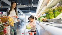 A mom and daughter are walking side by side in the produce aisle of a grocery store, pushing a regular and kid-sized shopping cart.
