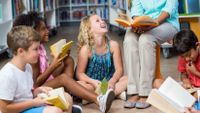 A group of young students sit on the floor around their teacher’s legs as she reads to them.