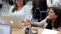Smiling teacher crouched down speaking with a smiling student who's working on her laptop