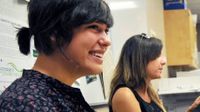 Two young women are sitting in a classroom, one smiling widely.
