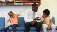 An adult male in a white button up shirt, tie, and black pants is sitting between two young boys in chairs against the wall of an administrative office. He's talking to one of them, while the other boy has his hands over his ears.
