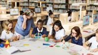 A group of elementary students sit at a table analyzing geometric shapes as their teacher assists.