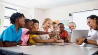 A group of elementary school aged children sit at a table talking with an adult and looking at a tablet screen together.