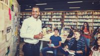 A smiling teacher stands in front of a whiteboard collecting ideas from several seated students.