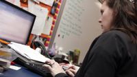A side profile, closeup of a young, female teacher typing while looking at her laptop. She's sitting in her classroom toward the front of the room next to a whiteboard.