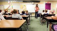 Elementary students are sitting at their desks, looking at the front of the class towards their teacher.