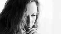 A closeup of a young teenage woman's face. She's looking down and her chin is resting on her clasped hands. Her long, wavy hair is covering half of her face.