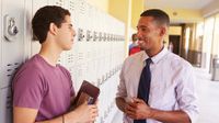 A male teacher and teenage boy are smiling, talking in a school hallway. The student is leaning against his locker, holding a folder and book.