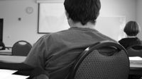 A black and white closeup of the back of a young man sitting at a long table towards the back of a classroom. He's looking toward the front of the classroom, and there's one other student sitting two tables ahead of him to the right.