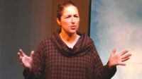 An adult woman with her hair pulled back in a ponytail is wearing a cowl neck maroon and beige checkered hoodie, standing with her elbows at her sides and her hands raised, as if she's addressing an audience.