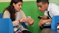 A teenage boy and girl are sitting across from each other in class talking to each other. The girl is pointing at her smart phone.
