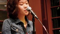 A young teenage girl with shoulder-length hair and a blue jean jacket is standing next to a microphone stand with the microphone up to her mouth. She's standing in the corner of a room with two glass windows behind her.