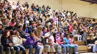 In the multipurpose room, a large group of middle school students fill one side of the bleachers, some clapping.