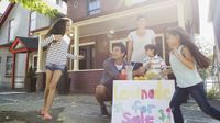 Two parents and three children are playing outside on the sidewalk in front of their house, setting up a lemonade stand.