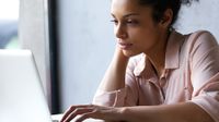 A closeup of a young adult female sitting at a desk, looking at her laptop. She has one hand on the mousepad, and the other is resting against her face with her elbow on the desk.