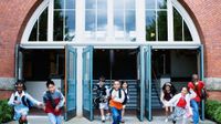 Eight elementary-age students are smiling, running out of the school building through three opened doors.