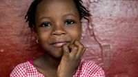 A closeup of a young girl, close-mouth smiling, with her chin resting on her right hand.
