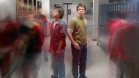 Two students are standing back to back in the middle of the school hallway. Other students are walking past them, blurred, as if to capture all the students walking through the hallway throughout the day.