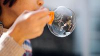A closeup of a young kid blowing a big bubble. He's to the left of the photo, the top of his head out of frame, and his arm is in front of his face holding an orange plastic device to blow bubbles.