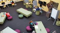 Teachers and students are sitting in a uniquely designed and furnished classroom with different sitting chairs and different sized and shaped tables.