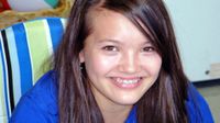 A closeup of a young girl, from the chest up, sitting in a striped white, green, and blue beach chair. She has long, brown hair, braces, and is wearing a blue polo shirt. She's smiling and looking directly at the camera.