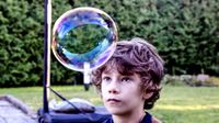 A young boy wearing a striped blue and orange t-shirt is looking at a bubble in front of his forehead that is almost the size of his head. He's outside. Behind him is grass next to a brick floor with a basketball hoop and a tree fence.