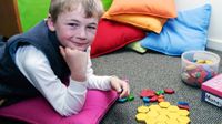 A young boy is lying on the floor, his elbows on a small, square pink pillow. He's looking at the camera with colorful shaped blocks in one hand, and more on the floor.