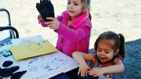 Two young girls are standing on grass next to a table with paper, stamp ink, crayons, and glove-like cloth that they're using as stamps. One girl is pushing her glove into the stamp ink, and the other girl is holding up her glove.