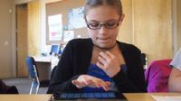 A young girl is sitting at a table in a classroom, working on a tablet.