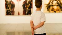 A young boy in a white t-shirt is standing in a museum, with his hands behind his back, looking at artwork.