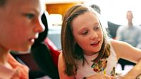 A young girl is sitting, looking at something in front of her and smiling. She has a yellow crayon in one hand and her other hand is palm down on a worksheet. A young boy is sitting a couple feet away from her at the same table, looking at the same thing.