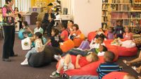 A group of about 20 young children are sitting and laying down on bean bags in a library. They're listening to a teacher standing in from of them and talking to them.