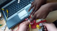 A closeup of two pairs of hands making something out of legos and tin foil over a desk, laptop, and red, yellow, and black wires.