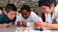 Three young boys are hovering over their classroom table, smiling. They're holding and looking at nuts, bolts, and screws that are lying on a piece of lamented paper with two circles drawn on it.
