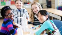 Four young students are smiling and sitting at two desks pushed together. Five paper-made windmills are on their desks.
