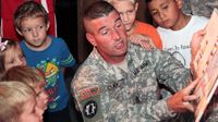 A U.S. army soldier in uniform is kneeling, reading to a group of young kids around him.