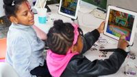 Two young girls are sitting in class with two tablets raised in front of them on their table. They're both looking at the same screen while one of them is playing a game on the tablet.