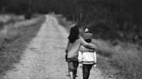 Two young kids are walking away from the camera along a gravel path with tall grass and trees on both sides. They're both wearing jeans and sweatshirts and one of them is wearing a beanie.