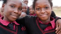 Two young girls are outside, smiling directly at the camera, hugging each other with one arm. They're both wearing black sweaters with red embroidered around the collar. Beneath their sweaters, they're wearing a pink, collared button up with a blue tie.